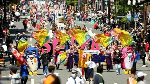 Толпа народа в разных цветых костюмах радостно идёт по проезжей части, играя на музыкальных инструментах