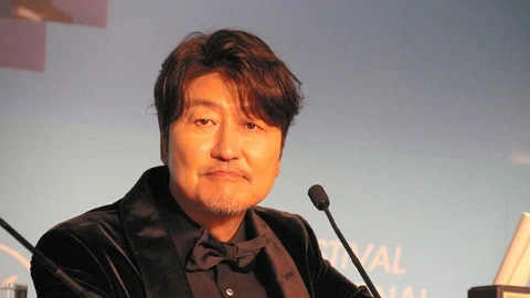 Мужчина азиатской внешности, с чёрными волосами и чёрными глазами, в чёрном пиджаке и тёмной рубашке, сидит за столом, рядом с микрофоном и сдержанно улыбается