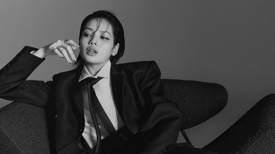 Девушка с тёмными волосами, в чёрном деловом костюме, в белой рубашке и чёрном галстуке, ехидно сидит на диване