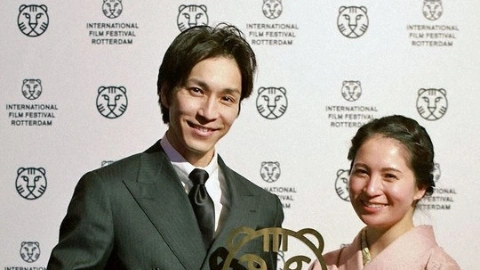 Мужчина с чёрными волосами и чёрными глазами, в деловом костюме, в пиджаке и галстуке, довольно улыбается, держа в руках статуэтку в виде тигра, рядом стоит женщина