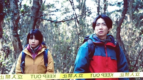 Мужчина с чёрными волосами, в сине-красной куртке, с рюкзаком за спиной, подозрительно смотрит вперёд, рядом стоит девушка с чёрными волосами, в коричневом пальто, с рюкзаком за спиной, оба стоят в лесу