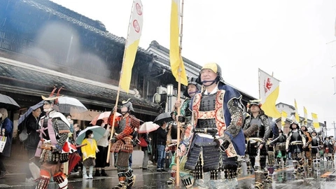 Пожилой мужчина в доспехах самурая идёт вдоль улицы, держа флаг, рядом идут другие такие же мужчины