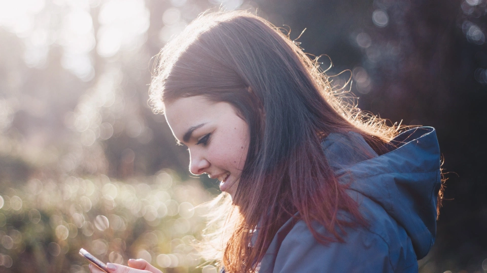 Девушка с каштановыми волосами, в синей кофте, находясь в лесу, радостно смотрит в экран телефона, который держит двумя руками