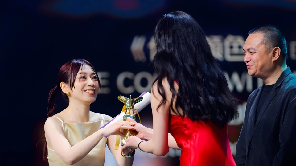 Женщина с коричневыми волосами, в бежевом платье, радостно получает статуэтку из рук высокой женщины с длинными чёрными волосами, в красном платье, рядом стоит мужчина