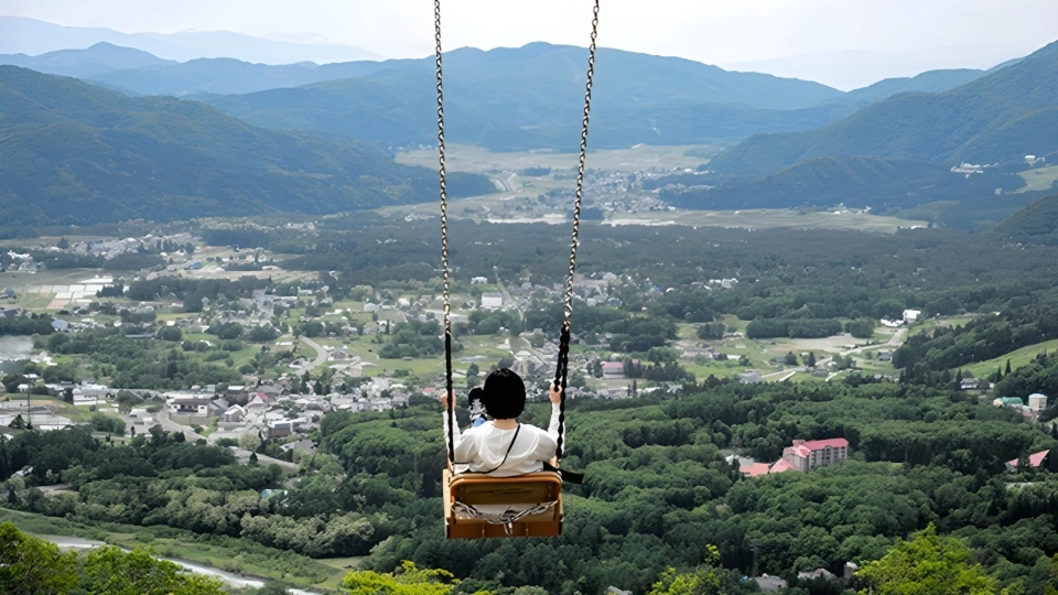 Юноша с чёрными волосами, в белой футболке, качается на качелях на фоне гор