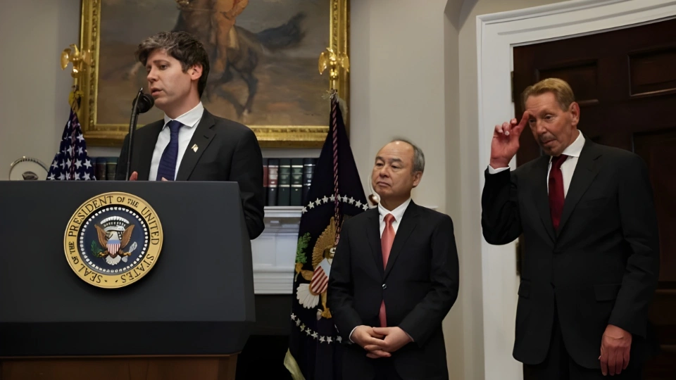 Мужчина в деловом костюме стоит за трибуной и говорит в микрофон, рядом стоят два других мужчины в деловых костюмах